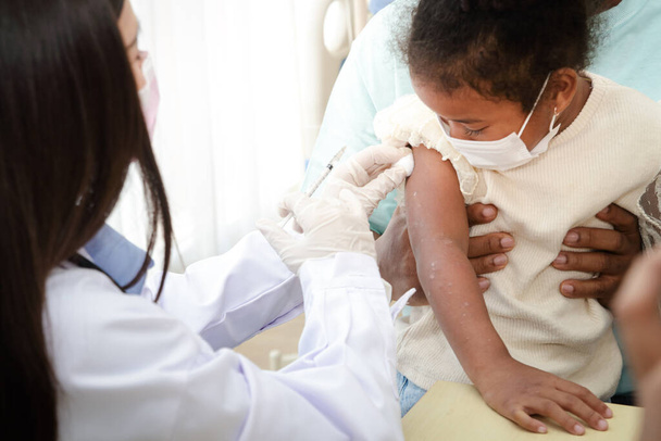 医師は若いアフリカ系アメリカ人の少女の腕の中にワクチンを注射し、免疫力を強化し、病気から守る。COVID-19ワクチンの概念。病院における医療サービス. - 写真・画像