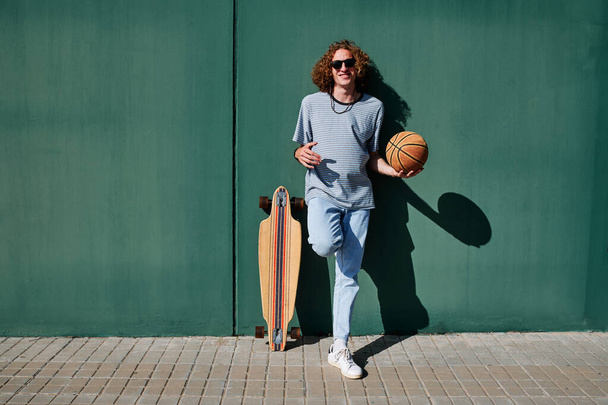 молодой человек в кудрявых волосах и солнцезащитных очках со скейтбордом и баскетбольным мячом с зеленой стеной позади него - Фото, изображение