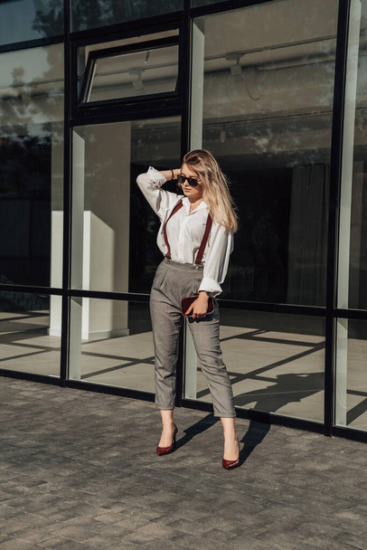 Vertical full length portrait of young small business owner standing near glass building. Business woman wear suit, sunglasses, suspenders and looking away. Freelance female outside. - Foto, immagini
