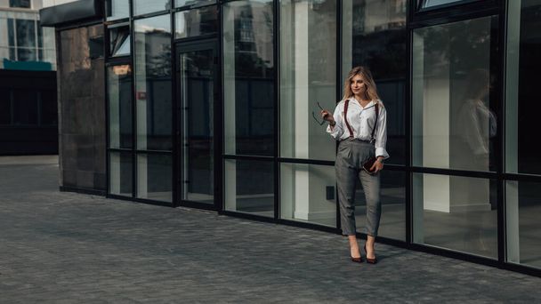 Full length portrait of young female CEO, manager, director, glass skyscraper. Business concept. Feminine gait. Gorgeous agent or entrepreneur holding sunglasses and clutch bag. Copy space. - Фото, зображення
