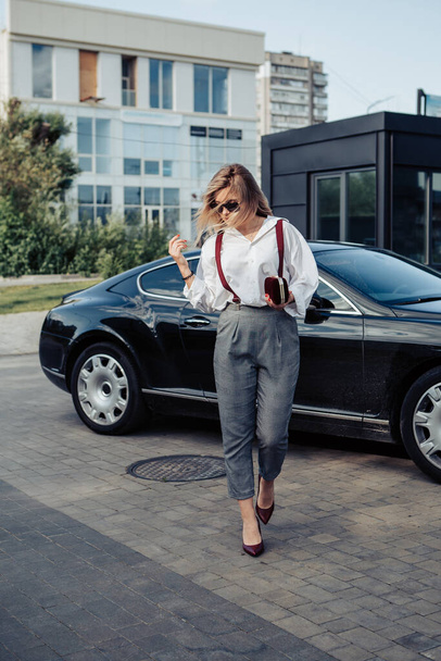 Porträt einer jungen Geschäftsfrau, die neben einem teuren Luxusauto mit Clutchtasche steht, mit weißem Hemd, Hose, Hosenträgern bekleidet und wegschaut. Bild mit Kopierraum. - Foto, Bild