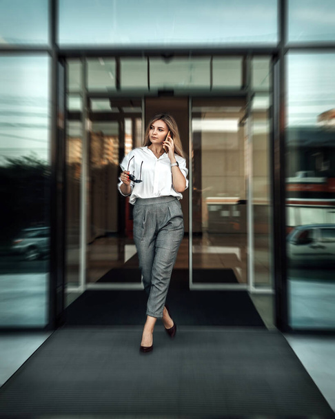 Vertical portrait of young woman small business owner comes out of an automatically opening door. Lady dressed in white bright shirt and pants hold mobile cell phone call speaking with client.  - 写真・画像
