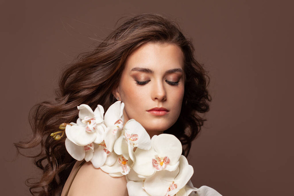 Aantrekkelijk vrouwelijk gezicht met heldere huid, gezond bruin haar, natuurlijke make-up en witte orchidee bloemen portret - Foto, afbeelding