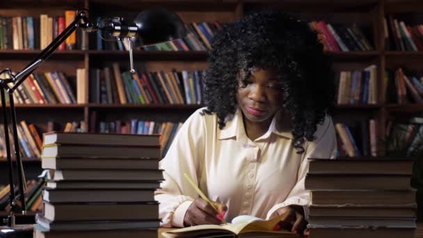 Mujer afroamericana escribe cerca de pilas de libros en la mesa - Imágenes, Vídeo