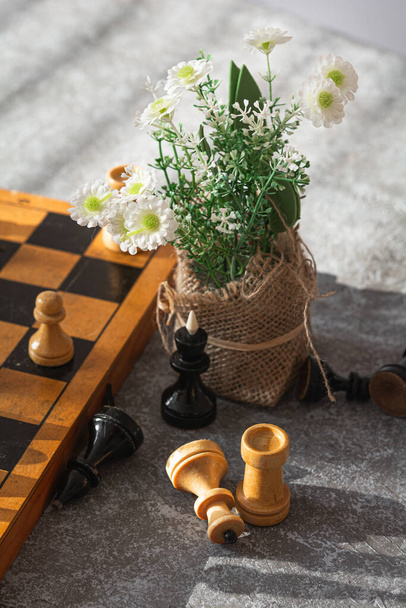 Шахматы. Шахматная доска, шахматные фигуры и цветы на серо-синем фоне в солнечный день - Фото, изображение
