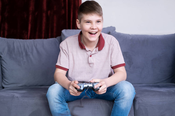 興奮した若い十代の男の子のゲーマーはジョイスティックでビデオゲームをプレイし、自宅でソファに座って. - 写真・画像