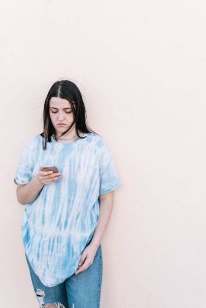 Teen girl in a tie dye shirt on a pinched orange wall looking at the smartphone - Φωτογραφία, εικόνα