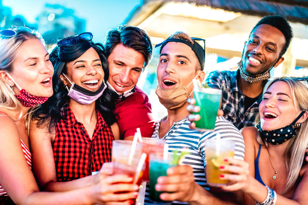 Happy multicultural people toasting at night bar with open face masks - New normal life style concept with milenial friends having fun together - Shallow depth of field with focus on middle guy - Photo, Image