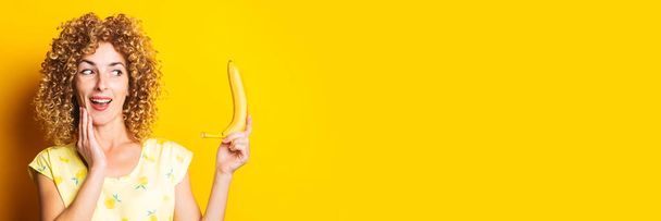 kudrnatá mladá žena vypadá překvapeně, když drží žlutý banán na žlutém pozadí. Nápis - Fotografie, Obrázek
