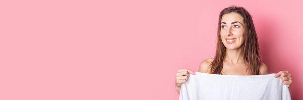Χαμογελώντας νεαρή γυναίκα μετά από ένα ντους καλύπτει τον εαυτό της με μια πετσέτα σε ένα ροζ φόντο. Μπάνερ - Φωτογραφία, εικόνα