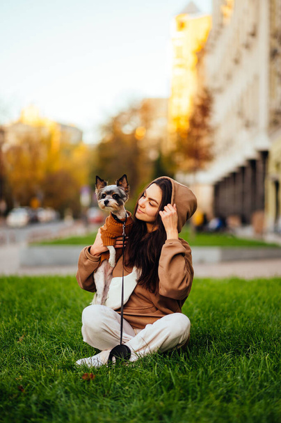 Függőleges felvétel egy divatos kutyatulajdonos nőről, aki a kis yorkshire-i terrierjét ölelgeti, divatos kapucnis pulóvert visel, a zöld füvön ül a városi utcában.. - Fotó, kép
