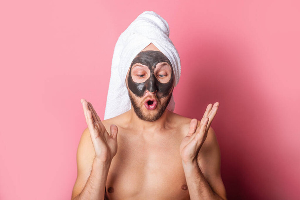 Ο νεαρός άνδρας κοιτάζει κατάπληκτος σε μια καλλυντική μάσκα στο πρόσωπό του σε ροζ φόντο. Έννοια φροντίδας δέρματος - Φωτογραφία, εικόνα