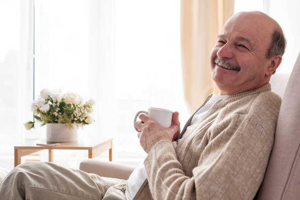 Портрет расслабленного пожилого человека, сидящего на диване с теплым напитком - Фото, изображение
