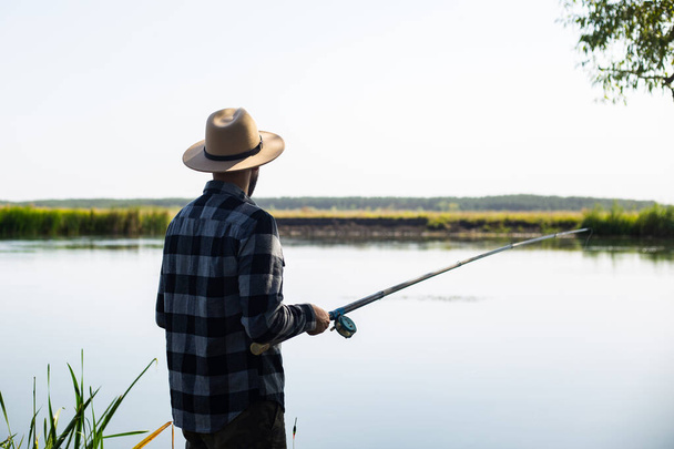 Homem com um chapéu e uma camisa xadrez está pescando em uma linha junto ao rio. - Foto, Imagem
