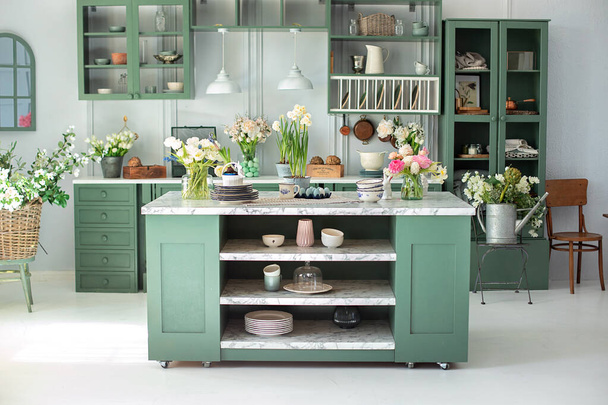 Zöld konyha belső bútorokkal. Stílusos konyha virágokkal a vázában. Fa konyha a tavaszi dekorációban. Otthoni dekoráció. Konyhai eszközök, edények és tányér az asztalon. konyha sziget étkezőben - Fotó, kép