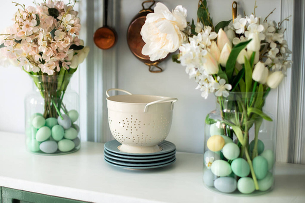 Zománcozott fehér színezék, tányérok, csokor virág vázában és húsvéti tojás a konyhaasztalon. Előnézet konyhapult konyhai eszközökkel, evőeszközökkel. Konyha szervezet. Tavaszi konyha belső dekoráció - Fotó, kép