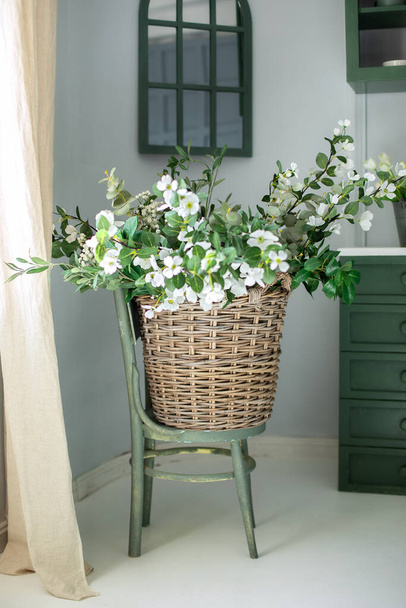 Branches of a blossoming apple tree in wicker basket in kitchen. Big basket with bouquet white flowers on wooden chair near wall. Beautiful bouquet of spring flowers in a basket.  Gift, mother's day - Photo, Image