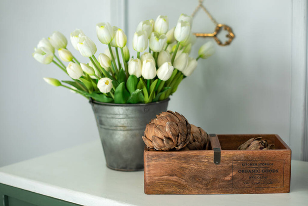 Samenstelling van gedroogde artisjok bloemen in houten doos en een boeket witte tulpen in een vaas als huisdecoratie. Moderne stijl bloemen arrangement in huis. Decor van droge bloemen thuis. Moederdag - Foto, afbeelding