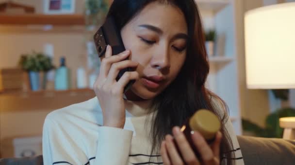 Beteg fiatal ázsiai nő tartja gyógyszert ül a kanapén használja okos telefon, hogy konzultáljon orvossal otthon éjszaka. Lány gyógyszert után orvosi rendelés, karantén otthon, Társadalmi távolság koronavírus koncepció. - Felvétel, videó