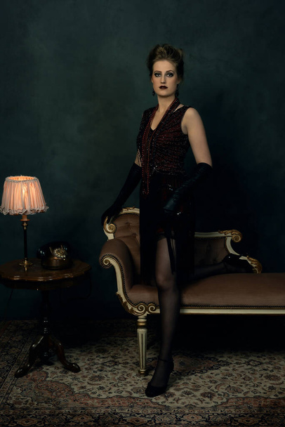 Спокуслива молода жінка в ретро-будуарному вбранні з намистами стоїть біля антикварного дивана поруч з журнальним столиком з настільною лампою і телефоном
. - Фото, зображення