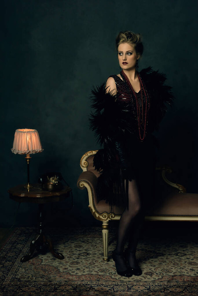 Viettelevä nuori nainen, jolla on musta boa retro boudoir-puvussa, jossa on kaulakorut antiikin sohvan vieressä sohvapöydän vieressä pöytälampulla ja puhelimella. - Valokuva, kuva