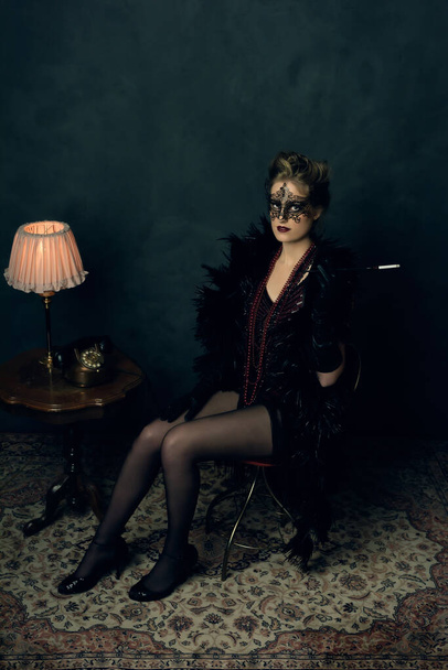 Mujer joven misteriosa con una boa negra y máscara en traje retro de tocador con collares sentados con un cigarrillo al lado de una mesa de café con una lámpara de mesa y teléfono. - Foto, imagen