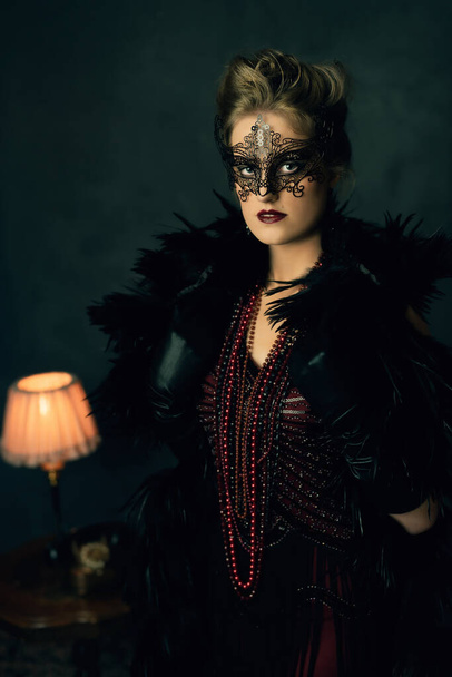 Mujer joven misteriosa con una boa negro y máscara en traje retro tocador con collares se encuentra delante de una mesa de café con una lámpara de mesa y teléfono. - Foto, imagen