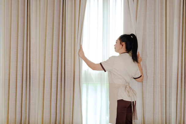 Fiatal szobalány behúzza a függönyt a hotelszobában - Fotó, kép