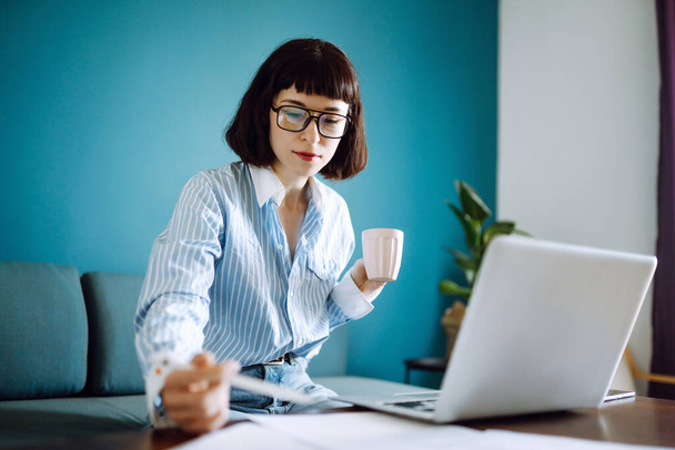 Beautiful  woman working on laptop computer while sitting at the living room, drinking coffee. Technology, freelance, shopping online, remote work, E learning concept.  - Фото, изображение