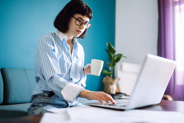 Beautiful  woman working on laptop computer while sitting at the living room, drinking coffee. Technology, freelance, shopping online, remote work, E learning concept.  - Фото, зображення