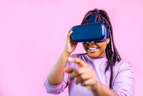 dark skinned woman sing glasses of virtual reality on pink studio background - Foto, imagen