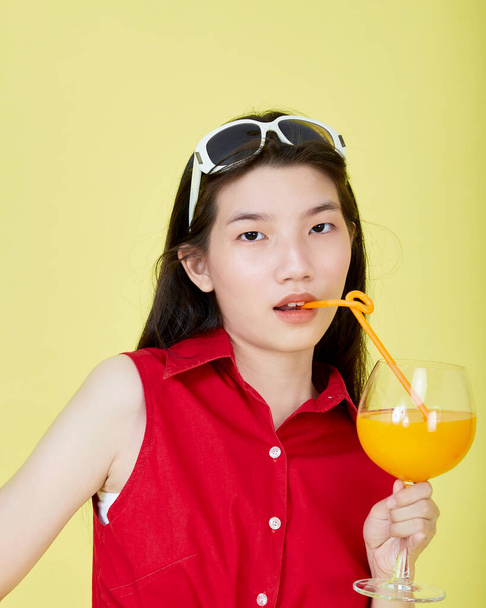 Όμορφη χαριτωμένο ευτυχισμένη ασιατική μαλλιά μελαχρινή γυναίκα το καλοκαίρι casual ρούχα με ένα ποτήρι φρέσκο χυμό φρούτων ποτό στούντιο πυροβόλησε απομονώνονται σε πολύχρωμο ένα κίτρινο φόντο - Φωτογραφία, εικόνα