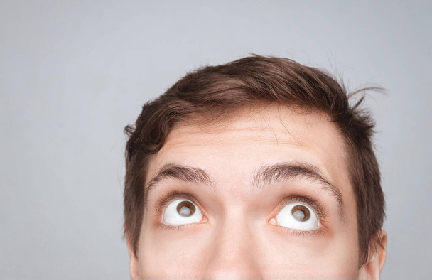 Man's eyes looking up on the light background. Empty place for a text or object. Close-up shot of shocked young man with round eyes, top half head. Eyes looking on top - Photo, Image