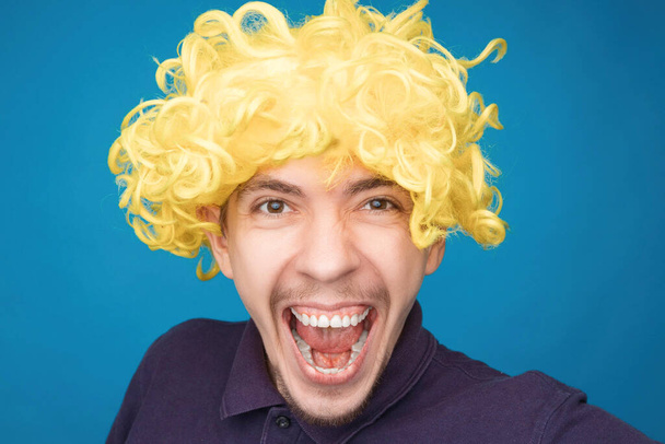 Mavi arka plandaki stüdyoda yüzünde mutluluk ifadesi olan bir adamın komik portresi. Peruk takan adam. Sarı saç. - Fotoğraf, Görsel