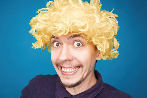 Mavi arka plandaki stüdyoda yüzünde mutluluk ifadesi olan bir adamın komik portresi. Peruk takan adam. Sarı saç. - Fotoğraf, Görsel