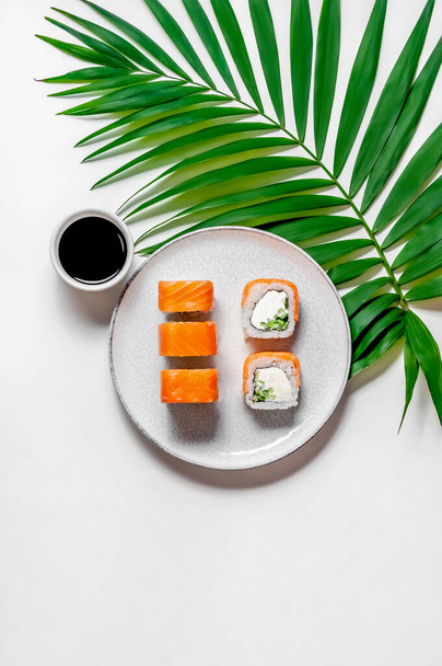 Філадельфійські булочки з рибою лосося, огірком і сиром на тарілці з соєвим соусом і пальмовим листям на білому тлі. Вигляд зверху лежав. Мінімальна концепція - Фото, зображення