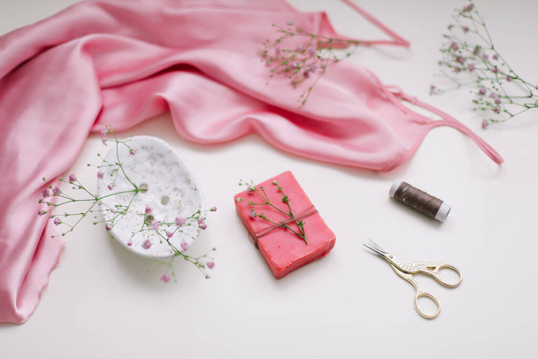 Overhead view of essential beauty items, top view of pink silk fabric, natural soap, flowers and decorative scissors, flat lay isolated on white background - Photo, Image