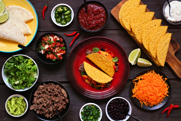 Taco bar table scene with a selection of ingredients. Above view on a dark wood banner background. Mexican food buffet. - Photo, Image