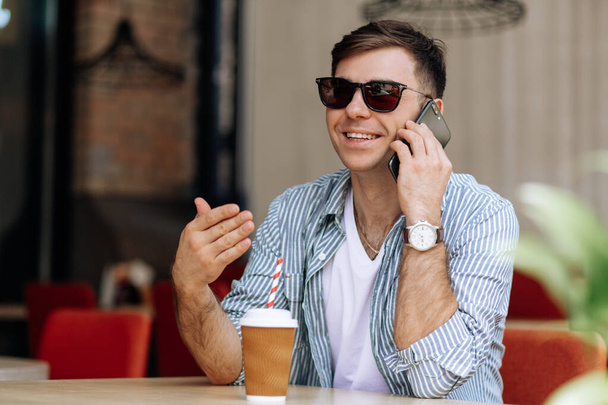 Schöner brünetter Mann mit Sonnenbrille sitzt in einem Café und telefoniert lächelnd - Foto, Bild