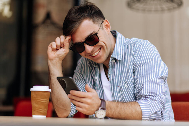 Handsome brunet man in sunglasses smiling into the phone while sitting in a cafe - Фото, изображение