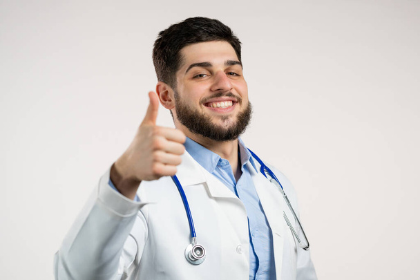 Doctor showing thumb up sign over white background. Positive young man in medical coat smiles to camera. Winner. Success. Body language. - Foto, afbeelding