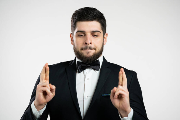 Young caucasian man praying with crossing fingers over white background. Guy in black tuxedo begging someone satisfy his desires, help with. - Fotografie, Obrázek