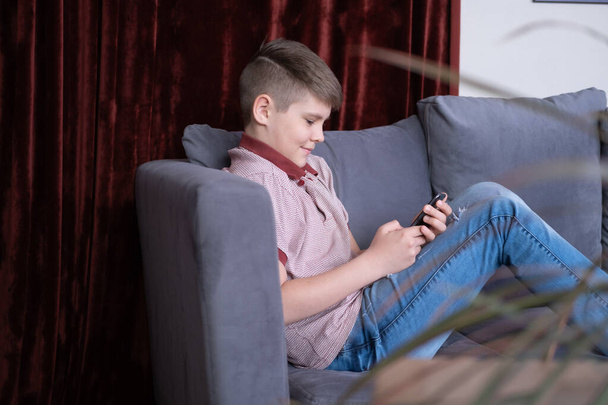 Joven adolescente guapo viendo contenido multimedia en un teléfono inteligente sentado en un sofá gris en casa - Foto, imagen