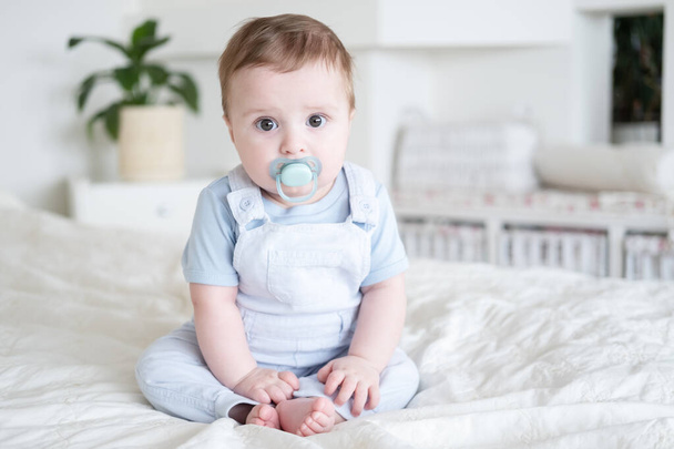 baby boy 6 months old with nipple in blu clothes and sitting on white bed at home. - Photo, Image