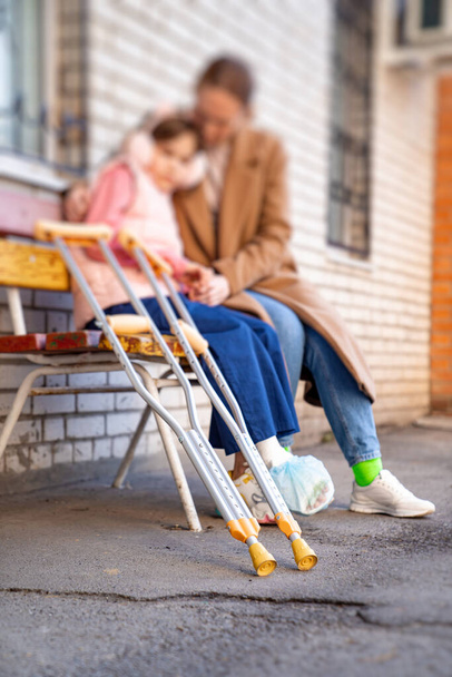mom with daughter with crutches sits on bench in yard of apartment building - Photo, image