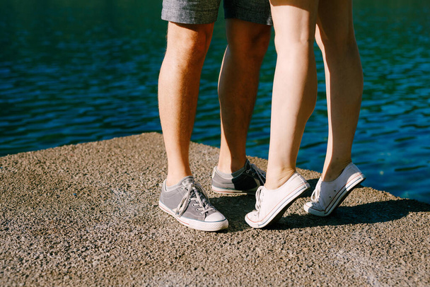 benen van een man en een vrouw in sneakers staan elkaar te knuffelen op de pier close-up, een vrouw staat op tenen  - Foto, afbeelding