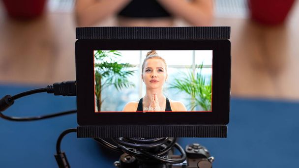 Profesjonalny aparat z zewnętrznym wyświetlaczem nagrywający młodą blondynkę w odzieży sportowej podczas medytacji na macie do jogi. Okna panoramiczne - Zdjęcie, obraz