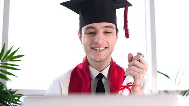 Счастливый молодой человек, говорящий и ждущий на онлайновом выпускном в своем ноутбуке со свитком в руках. Академическая одежда. Огромные окна и зелень. Социальная идея расстояния - Фото, изображение