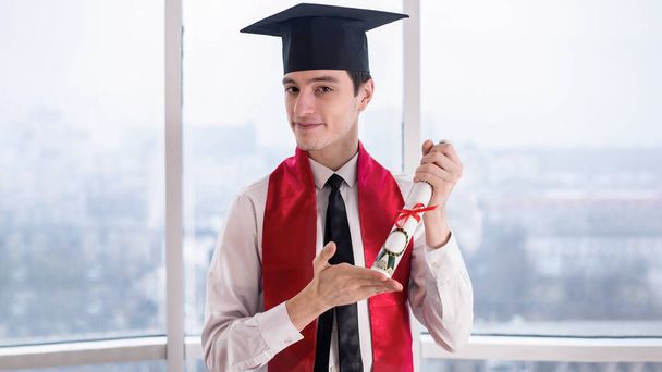 手にスクロールで自宅でオンライン大学卒業で幸せな若い男。アカデミックな服装。パノラマの窓。社会的距離の考え - 写真・画像