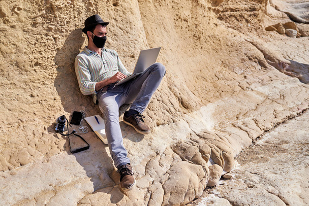 Um jovem hipster com um chapéu usa seu laptop na praia rochosa, ao lado dele ele tem um smartphone, uma câmera vintage e um bloco de notas - Foto, Imagem